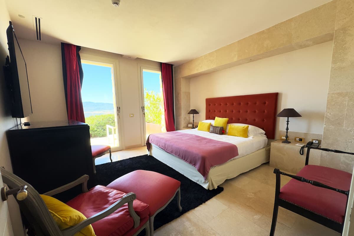 A hotel room with a bed, two chairs, a desk, a TV, and a view of the outdoors.
