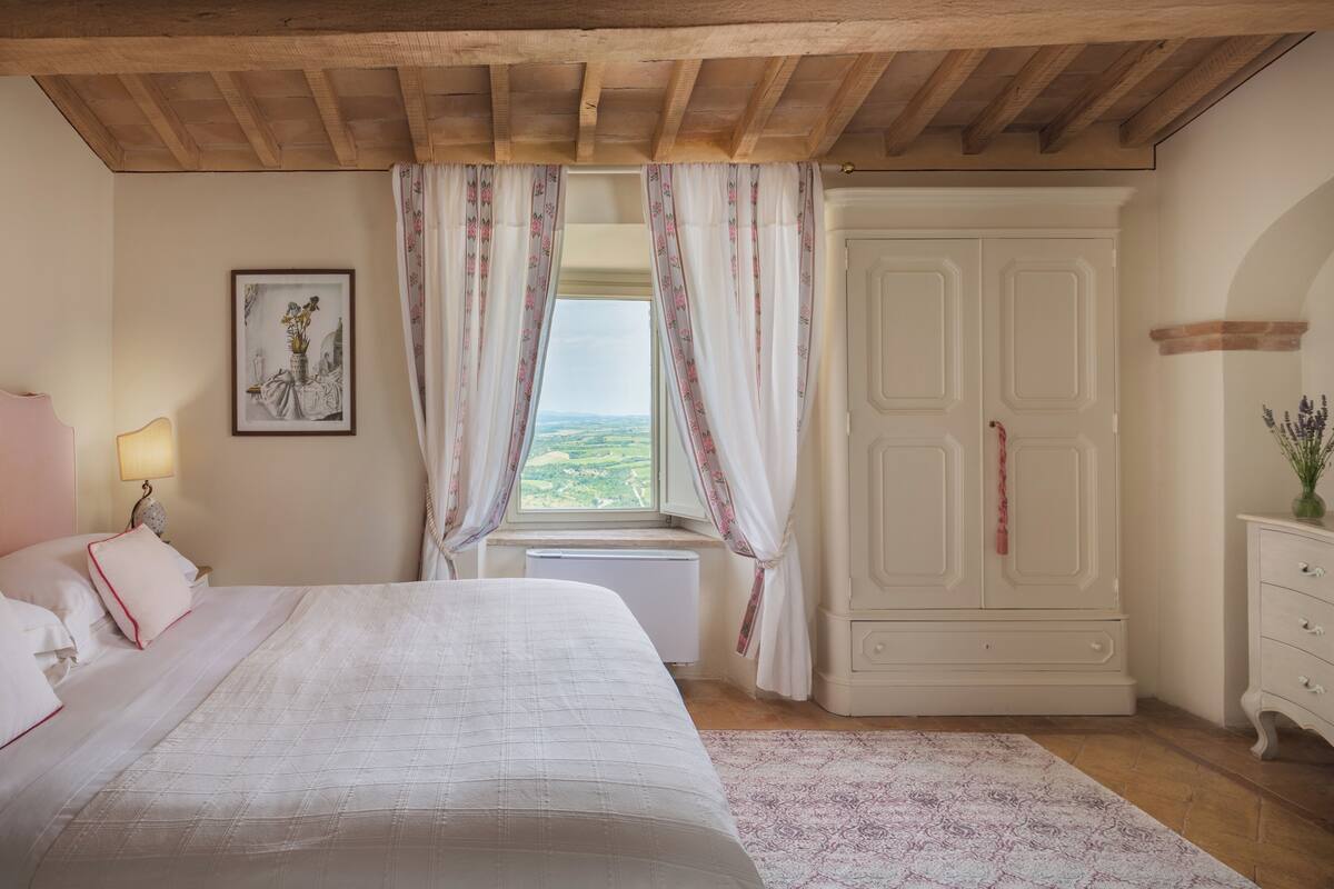 A bedroom with a bed, a window with curtains, a radiator, a wardrobe, and a lamp.