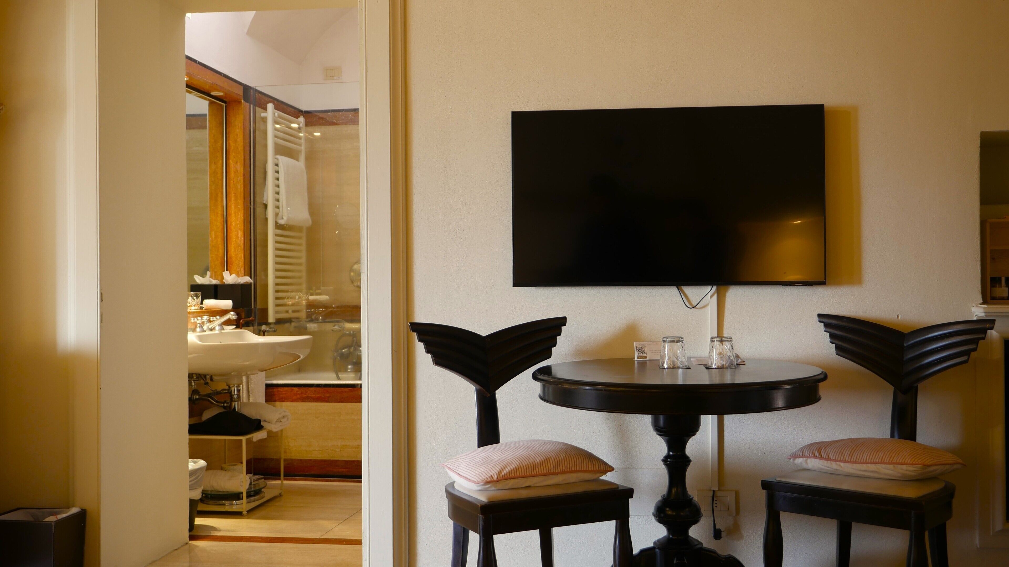 Une salle de bain équipée d’un lavabo, d’un miroir et d’un porte-serviettes. Un téléviseur est fixé au mur, au-dessus d’une table ronde accompagnée de deux chaises.