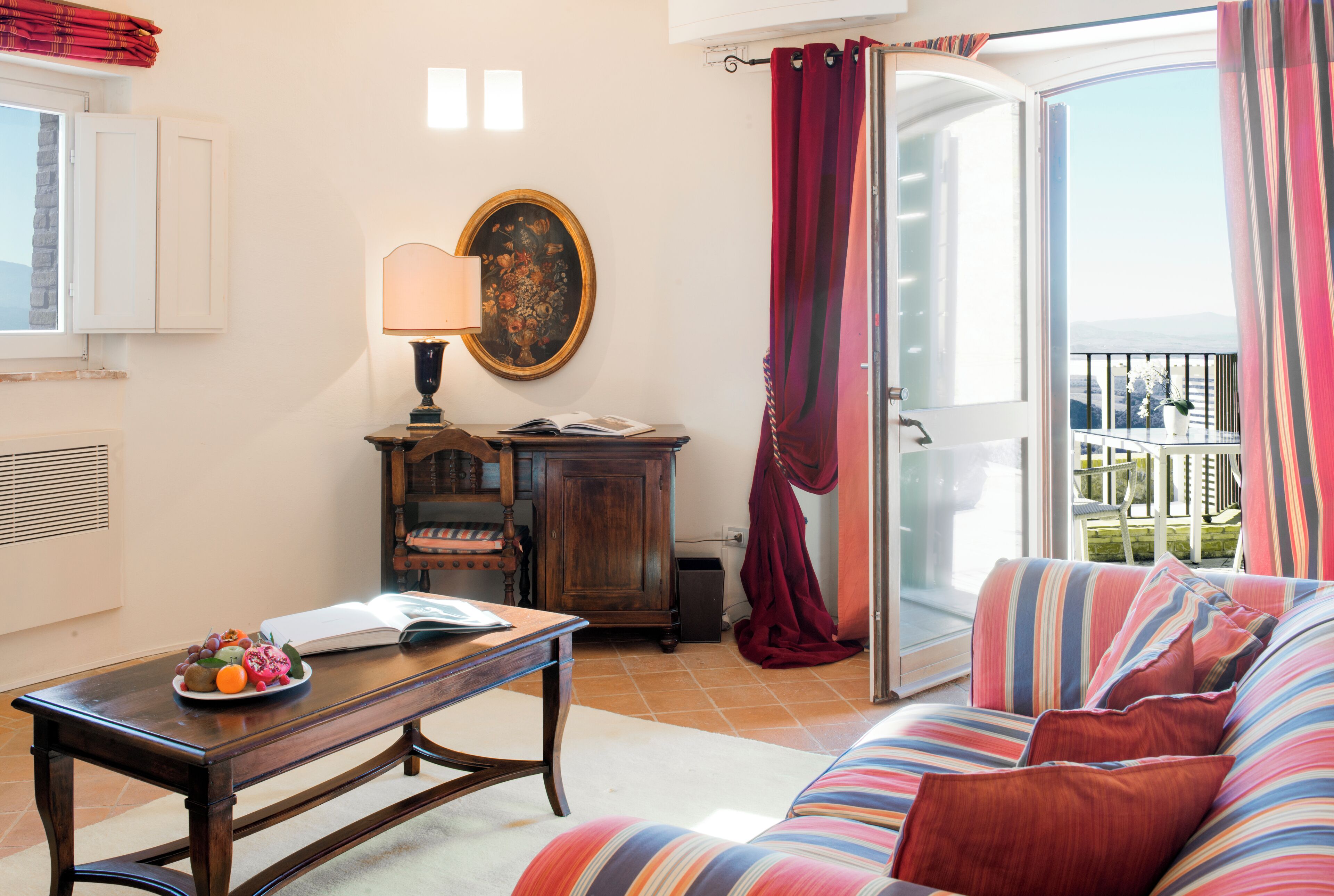 A living room with a sofa, a table with a book and fruit, a lamp, a painting, and a view through a sliding door to an outdoor balcony.