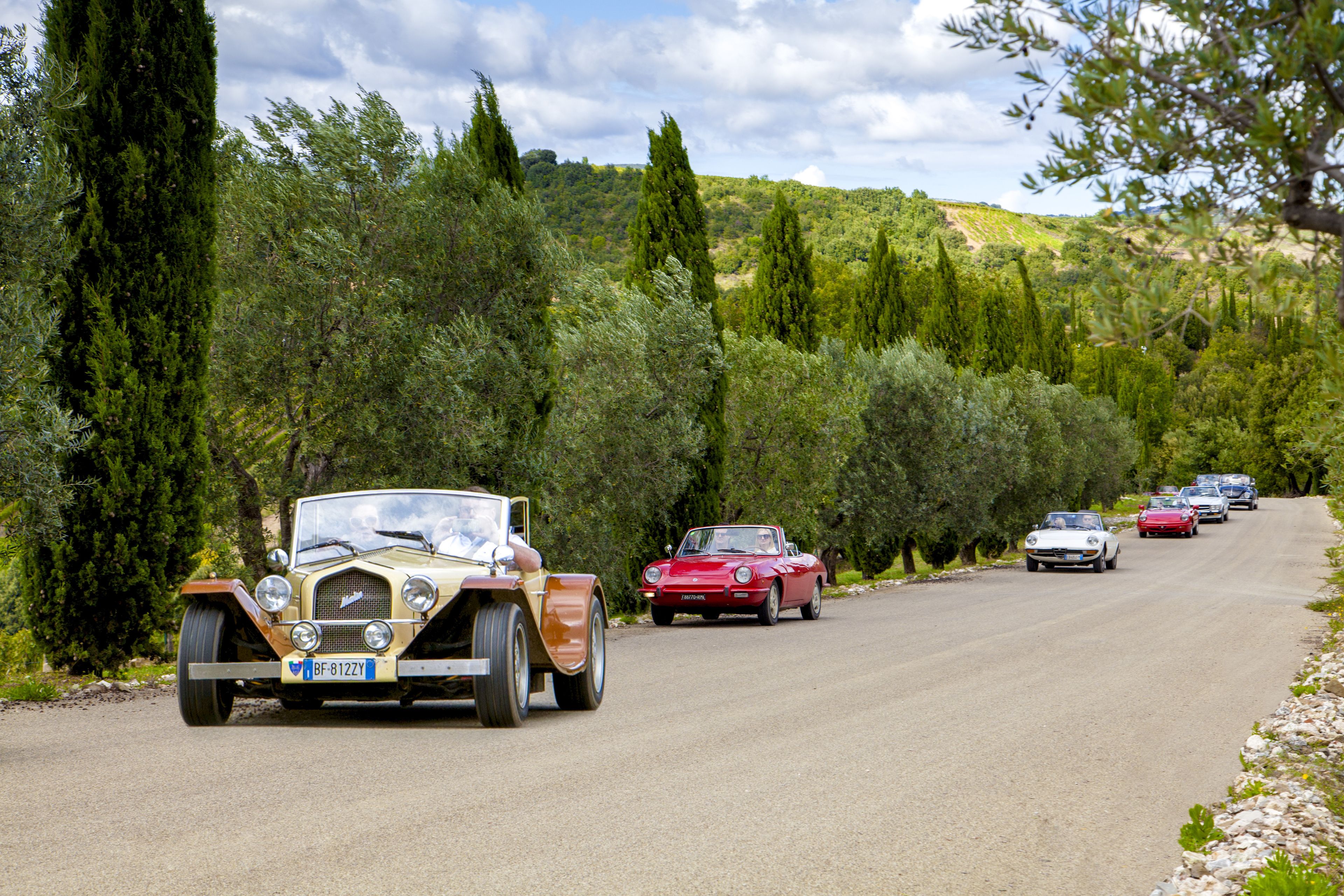 A vintage car driving on a road surrounded by trees and hills.