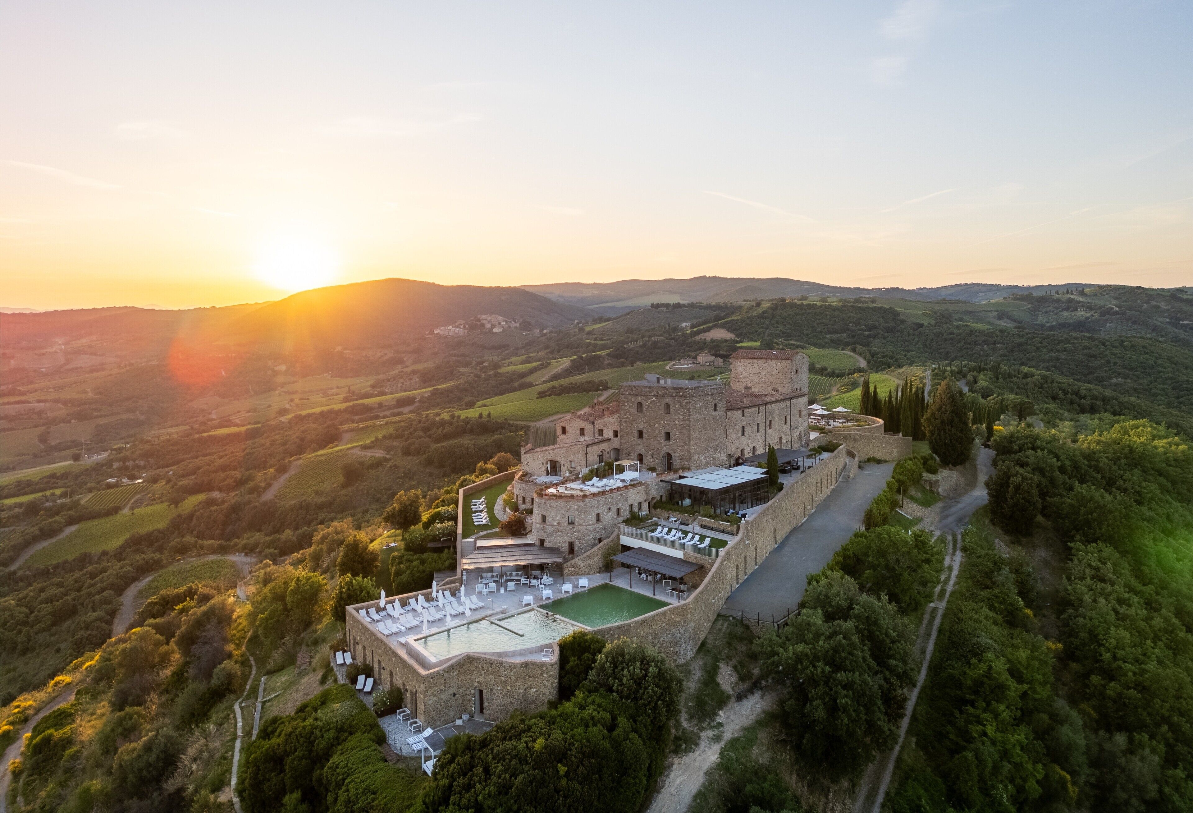 An aerial view of a castle with a swimming pool and outdoor seating area, surrounded by lush greenery and hills.