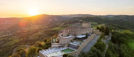 An aerial view of a castle with a swimming pool and outdoor seating area, surrounded by lush greenery and hills.