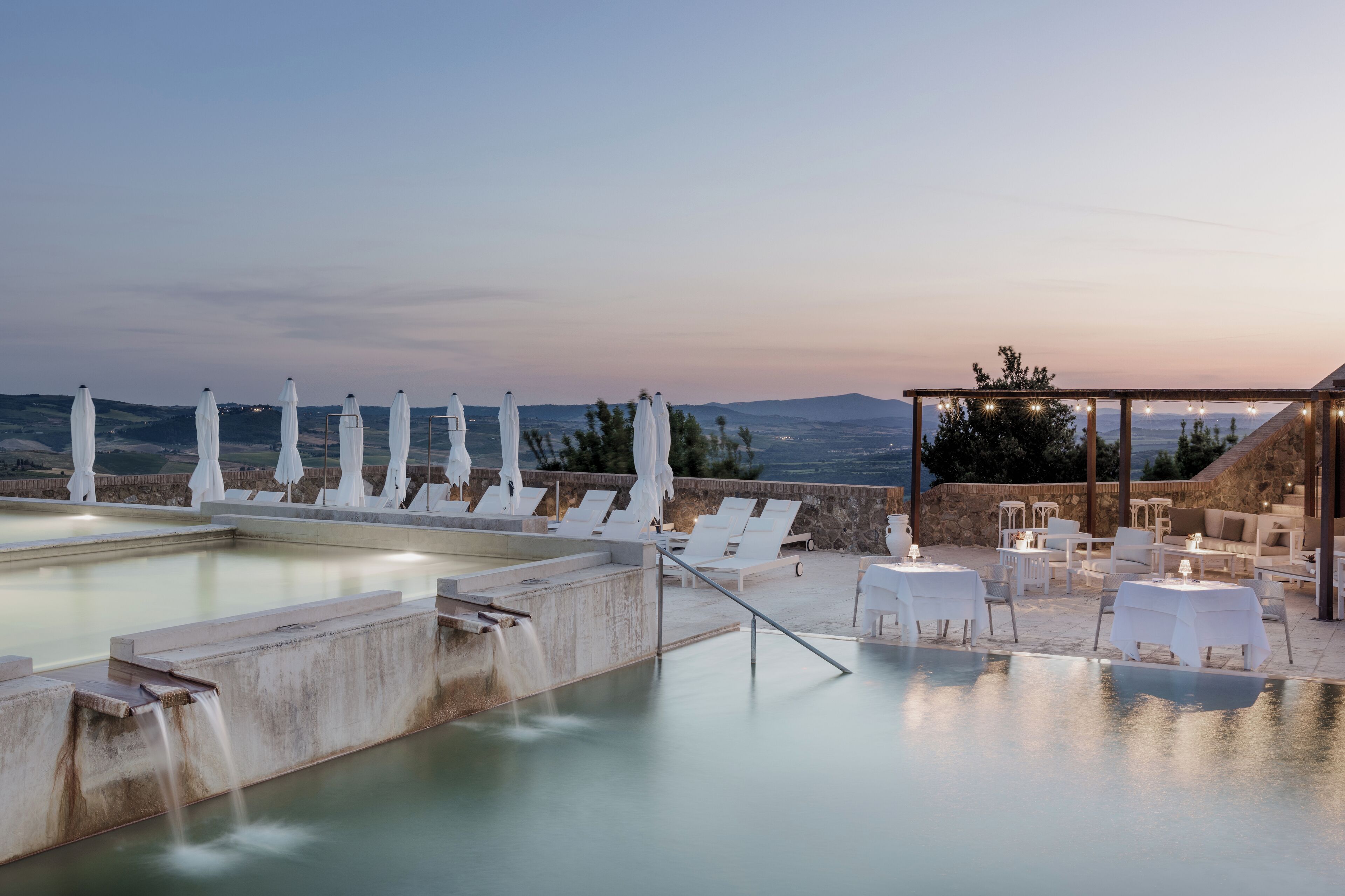Un área de piscina en la azotea con sillones blancos y sombrillas, un muro de piedra y vistas del paisaje.