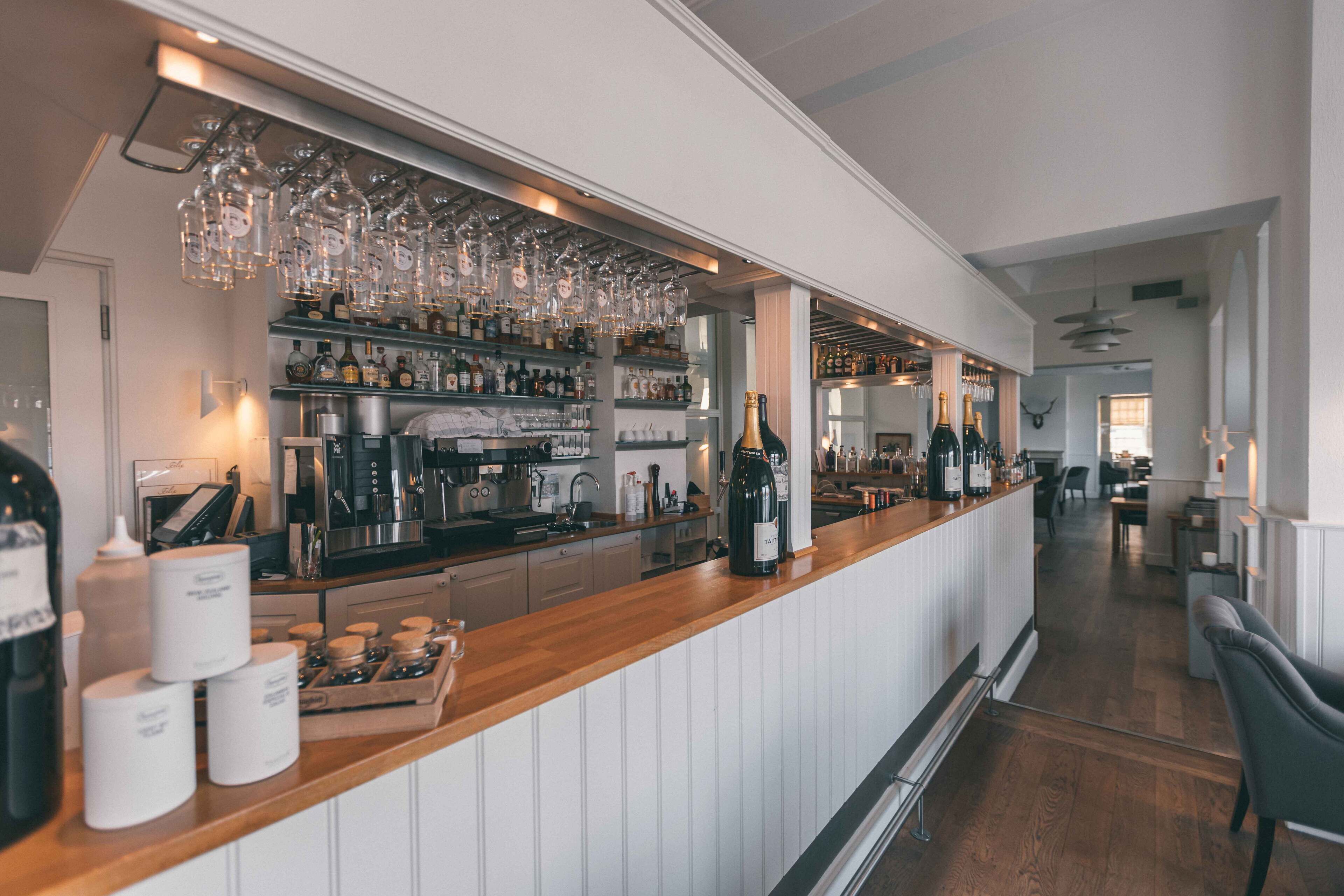 Una zona de bar con estantes llenos de botellas y copas, una barra de madera y vistas a un comedor.