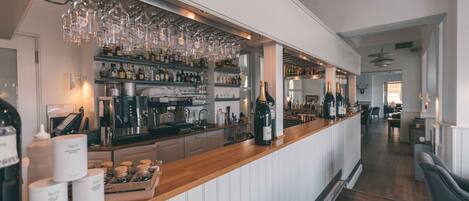 A bar area with shelves of bottles and glasses, a wooden counter, and a view into a dining area.