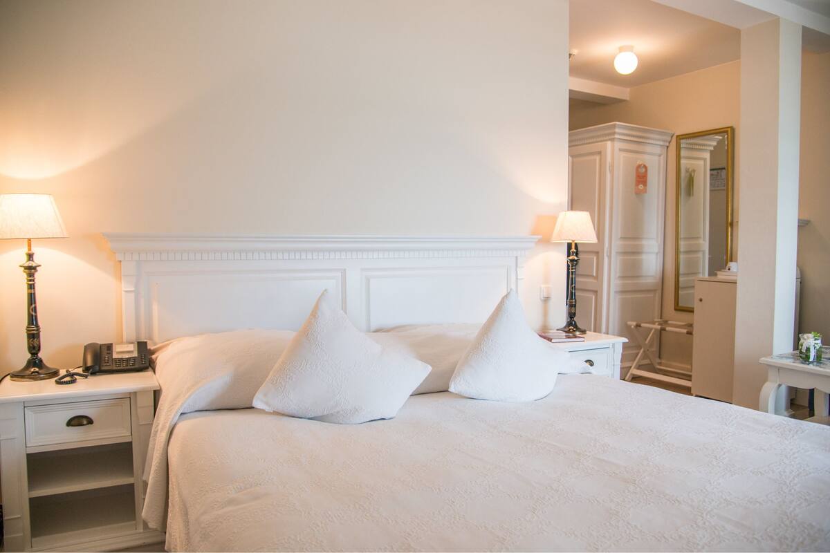 A neatly made bed with white linens and pillows, a bedside table with a lamp, and a mirror on the wall.