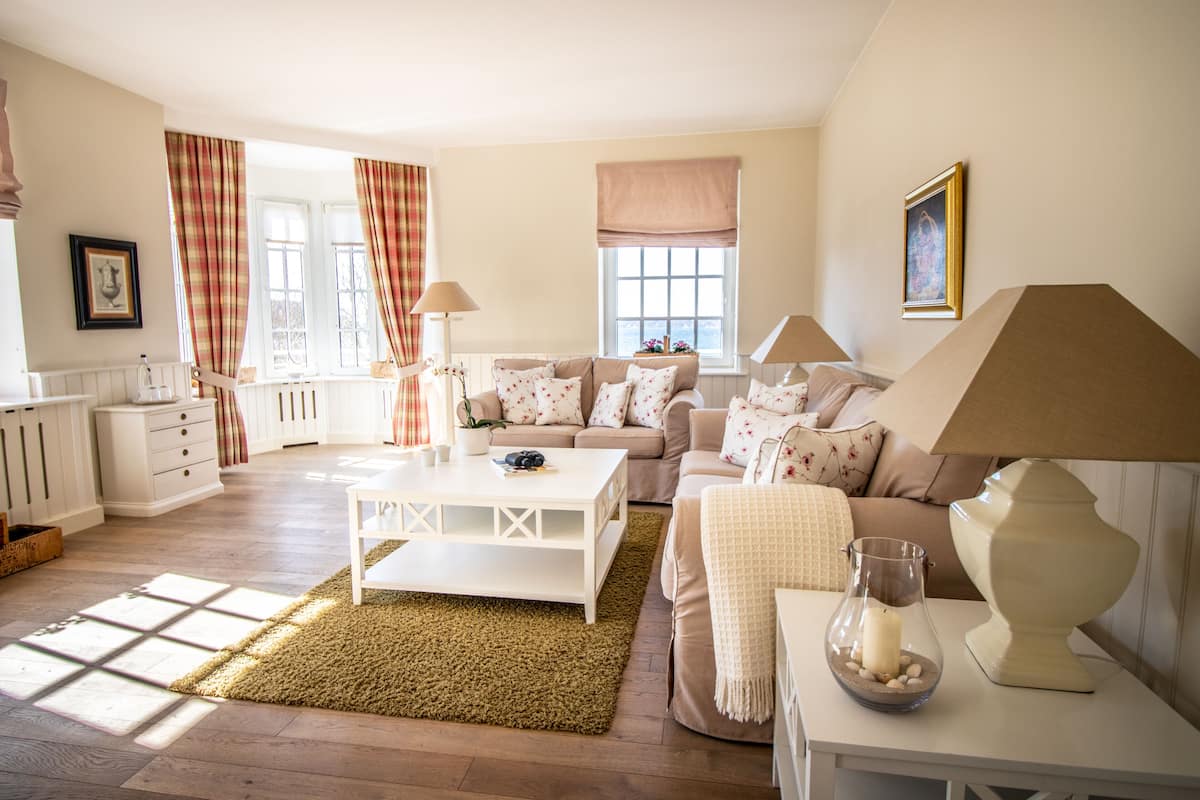A well-lit living room with a sofa, coffee table, and lamps. There are curtains on the windows, and a framed picture on the wall.