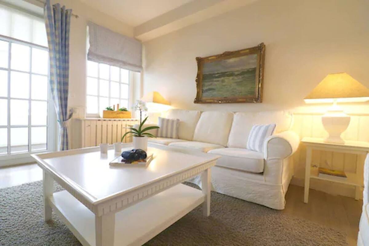 A white sofa, a white coffee table, and a lamp on a white nightstand in a room with a window and a painting on the wall.