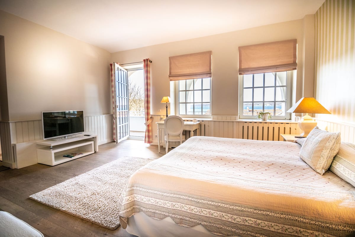 A bedroom with a large bed, a TV, a desk, and a chair. There are windows with curtains, a radiator, and a view of the outside.