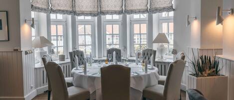 A dining area with a round table set for six, surrounded by six chairs. There are two lamps on either side of the table, and a potted plant is visible on the right side.