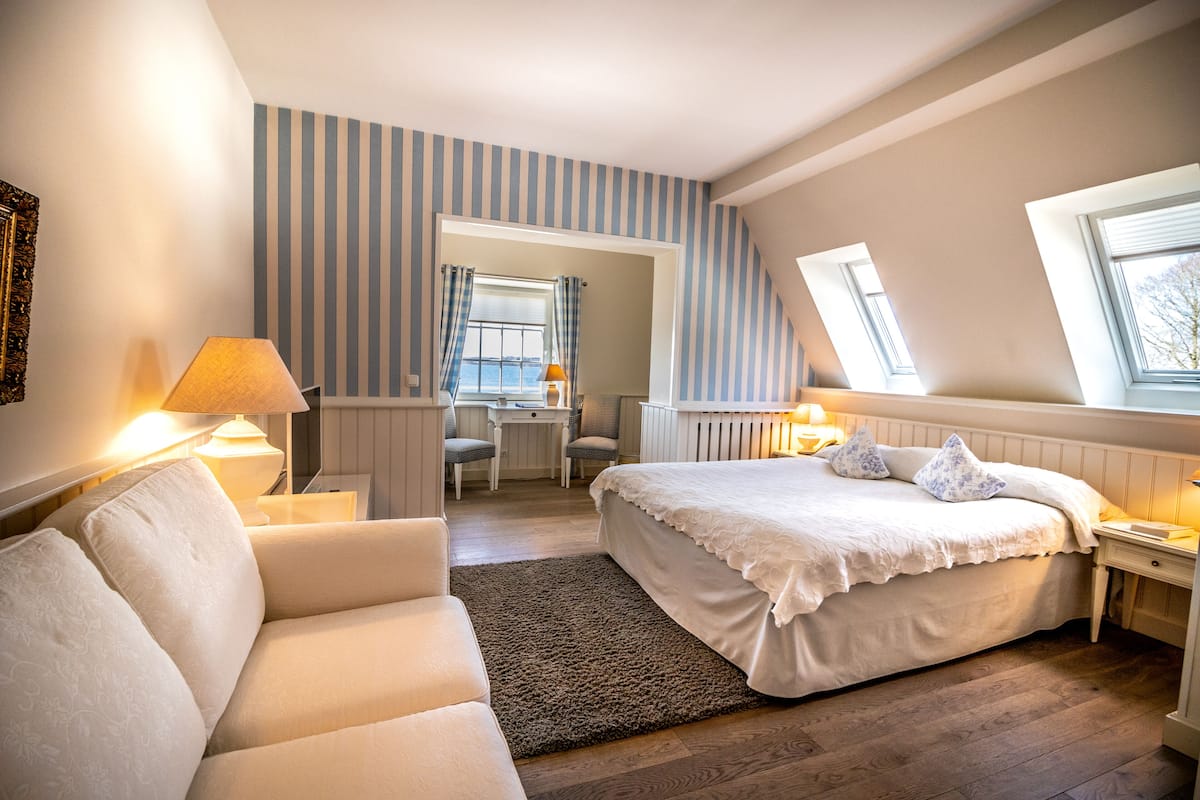 A bedroom with a large bed, a sitting area with a sofa, a desk, and a window with blinds.