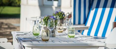A white outdoor table set with wine glasses and flowers, surrounded by white chairs and a blue-striped umbrella.