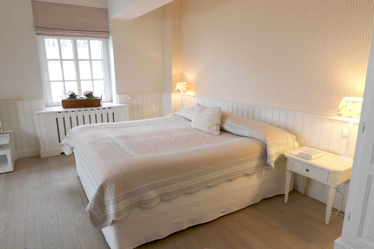 A bedroom with a large bed, white nightstands, a window with a view, and a radiator.