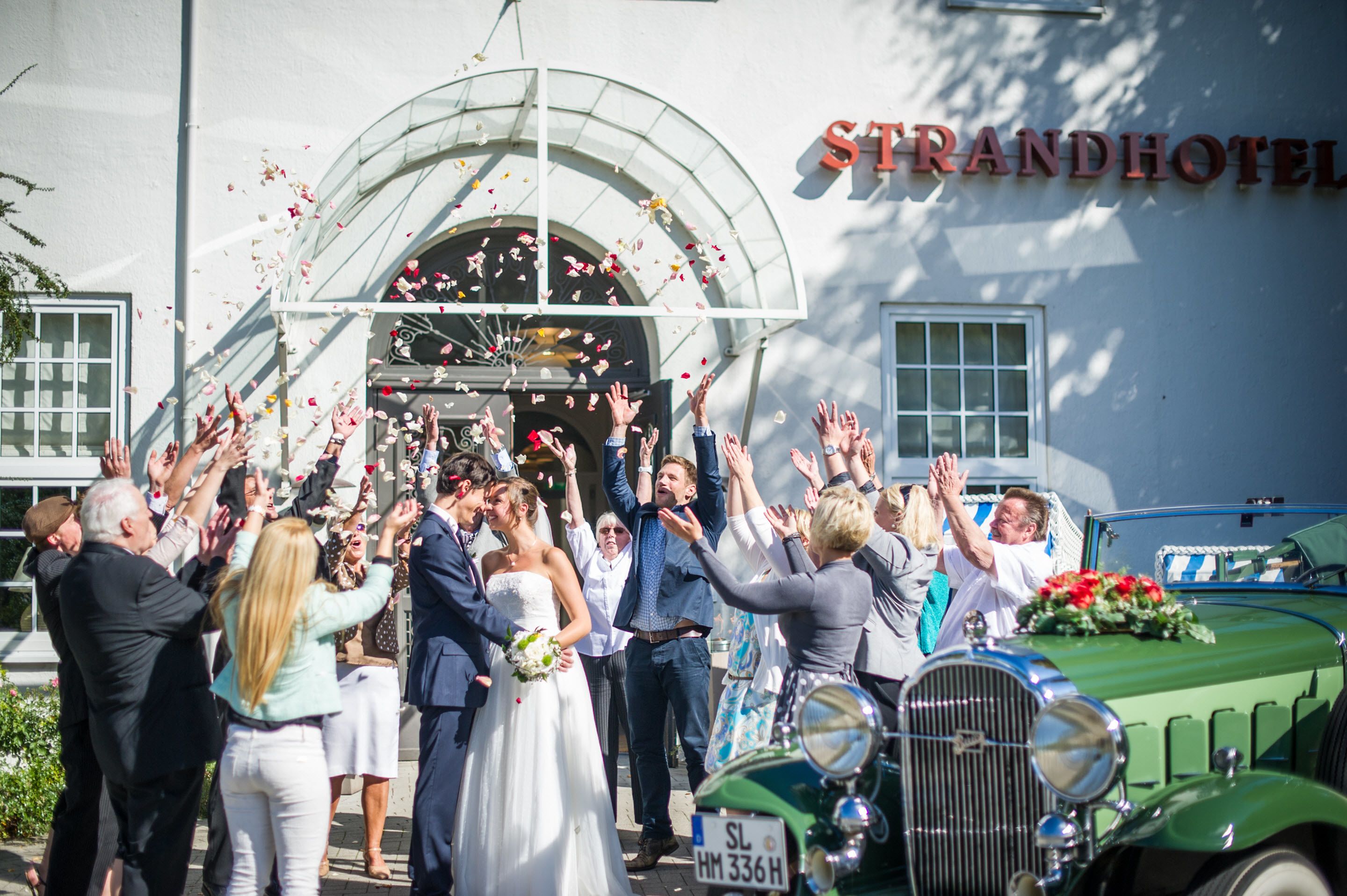 Eine Hochzeitszeremonie mit Gästen, die Rosenblätter werfen, ein altes, grünes Auto und ein Gebäude mit der Aufschrift "STRANDHOTEL".