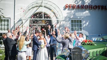Eine Hochzeitszeremonie mit Gästen, die Rosenblätter werfen, ein altes, grünes Auto und ein Gebäude mit der Aufschrift "STRANDHOTEL".