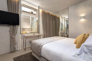 A modern hotel room with a large bed, a flat-screen TV mounted on the wall, a window with curtains, and a mirror reflecting the room.
