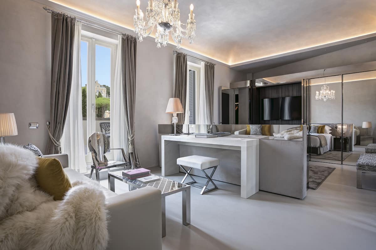 Un salon moderne avec un canapé blanc, une table basse en verre et un lustre. On y trouve une grande fenêtre avec des rideaux, un téléviseur et, au fond, on aperçoit un coin cuisine.