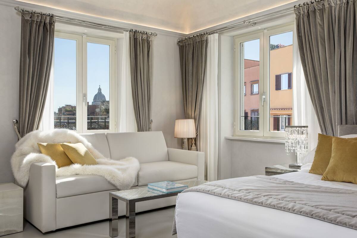 Une chambre d’hôtel avec un canapé blanc, un lit, une table de chevet avec une lampe, et une vue sur le paysage urbain à travers de grandes fenêtres.