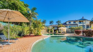 A swimming pool with lounge chairs and umbrellas, surrounded by lush greenery and a multi-story building in the background. - Aqualuna Beach Resort (Korora)