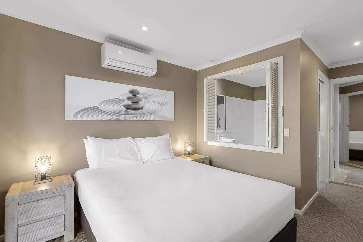A hotel room with a bed, a nightstand, a wall-mounted air conditioner, a large mirror, and a painting.