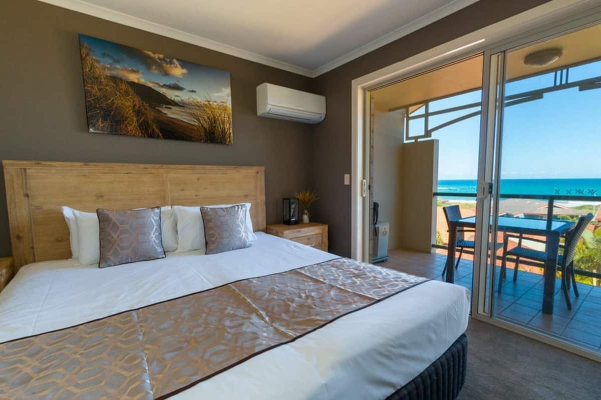A bedroom with a large bed, a painting on the wall, and a view of the ocean through a sliding door.