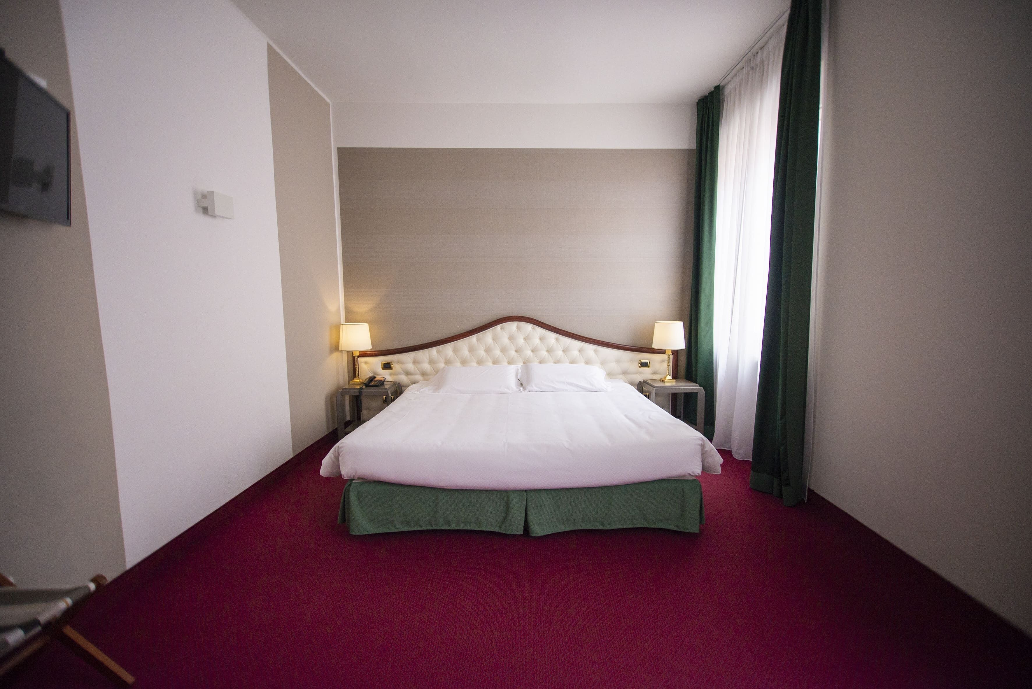 Habitación de hotel con una cama grande, falda para cama verde, dos lámparas de noche y una ventana con cortinas verdes.
