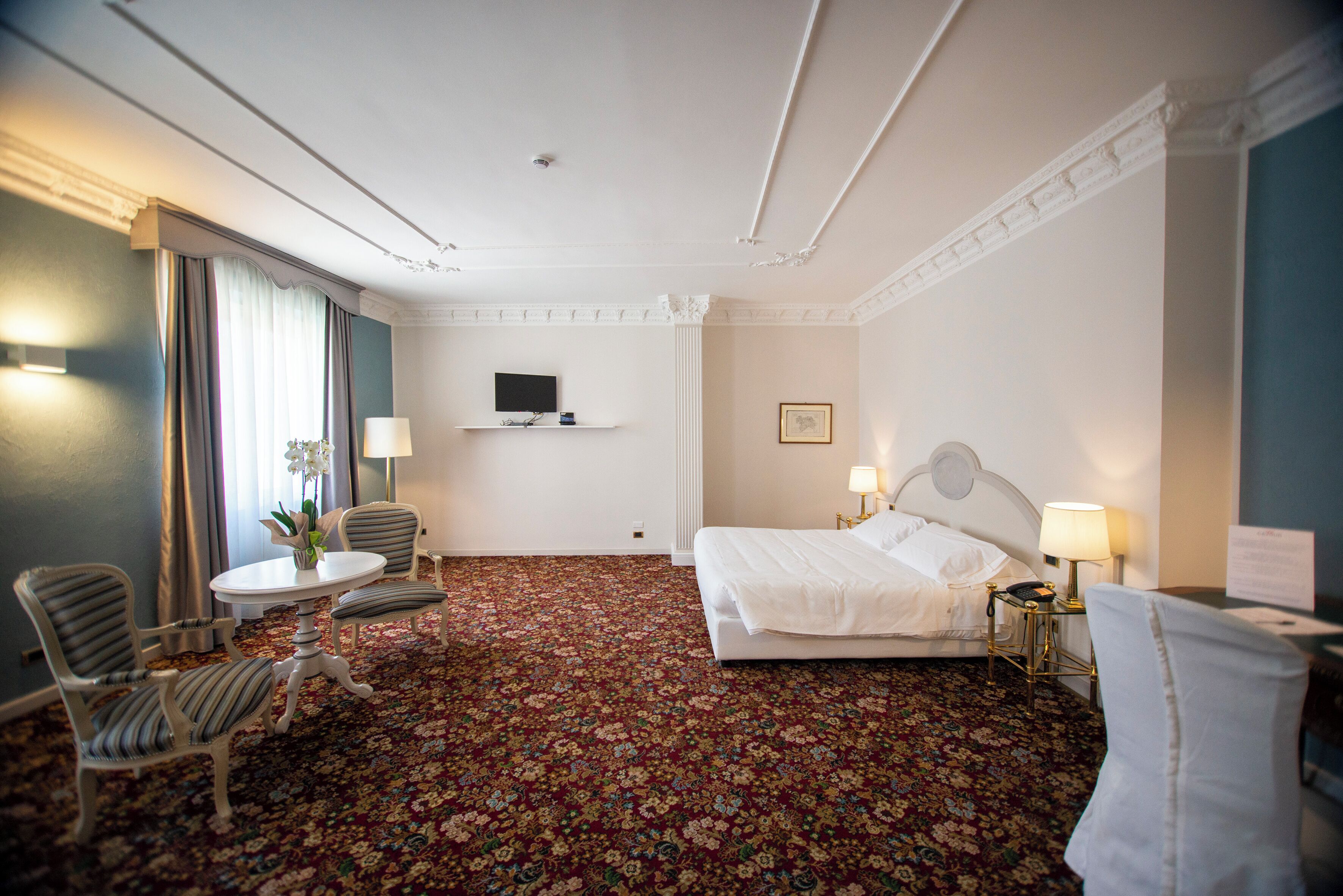 Une chambre d’hôtel avec un tapis à motifs floraux, un lit, un bureau avec une chaise, un téléviseur fixé au mur et une lampe sur la table de chevet.