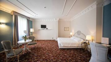 A hotel room with a floral carpet, a bed, a desk with a chair, a TV on the wall, and a lamp on the bedside table.