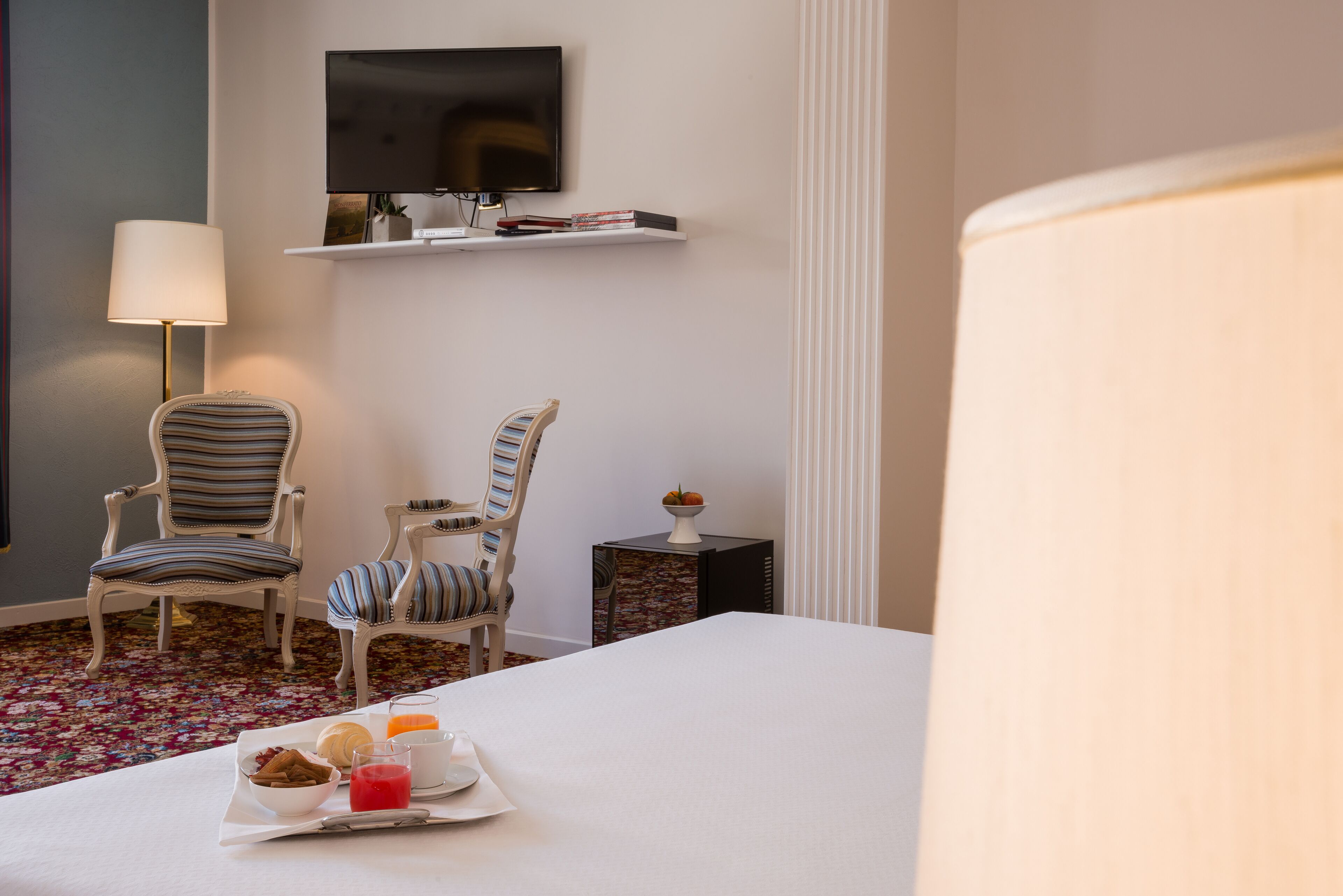 Una habitación de hotel con una cama, dos sillones a rayas, una mesita con una bandeja de desayuno, un televisor en la pared y una lámpara.