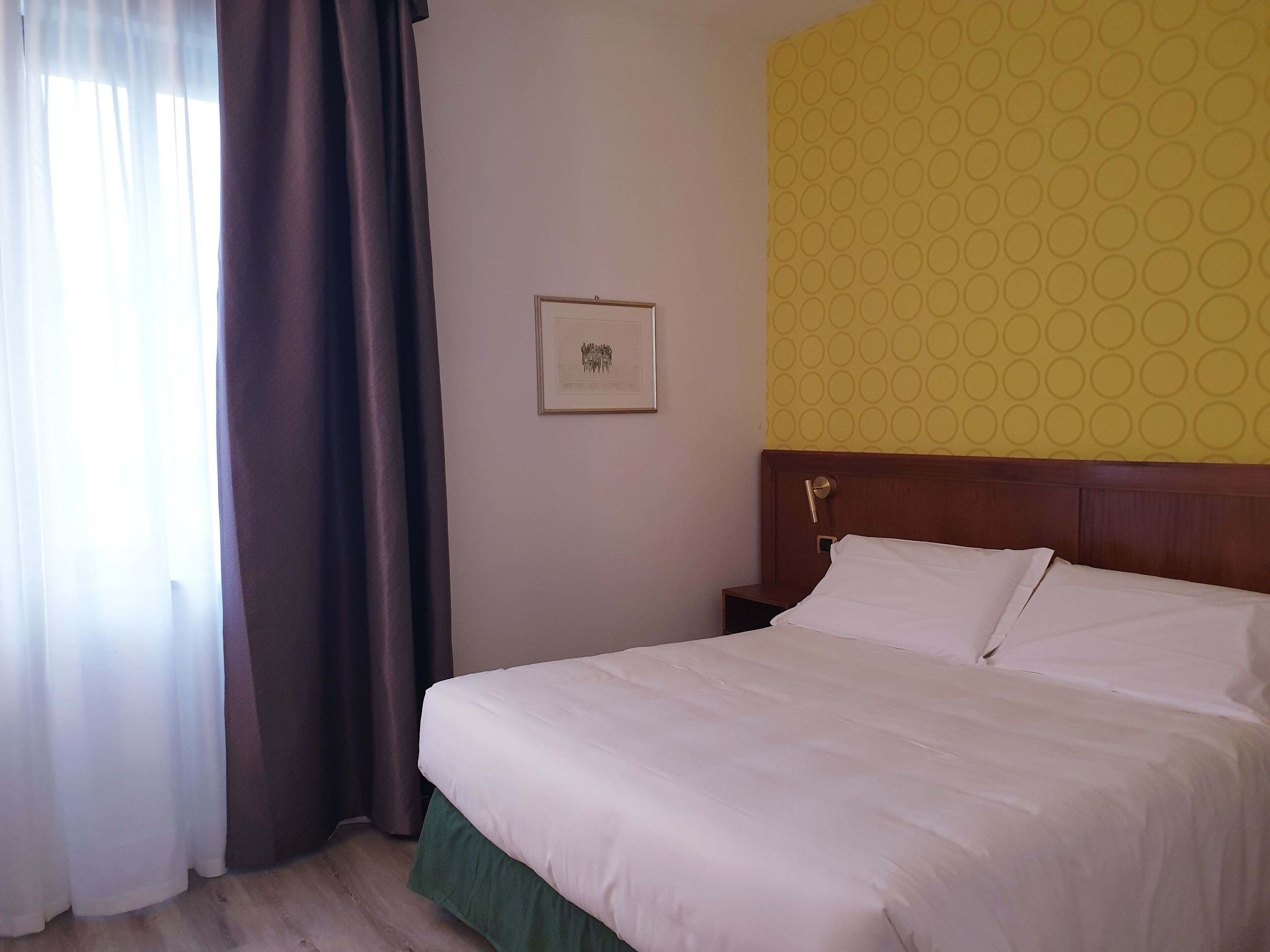 Ein Hotelzimmer mit Bett, Nachttisch, einem gerahmten Bild an der Wand und einem Fenster mit Vorhängen.