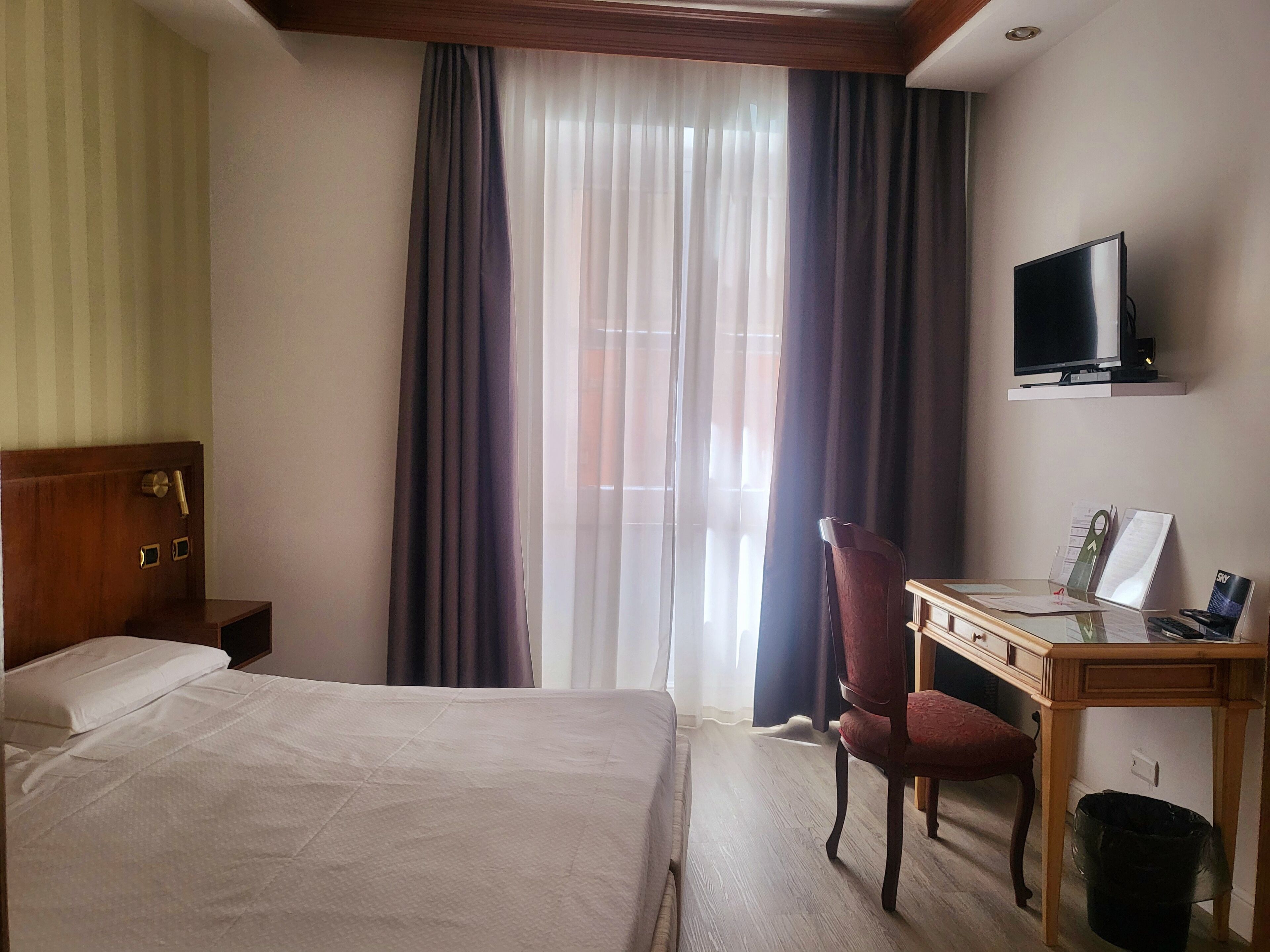 Una habitació d'hotel amb un llit, un escriptori amb una cadira, una televisió i una finestra amb cortines.