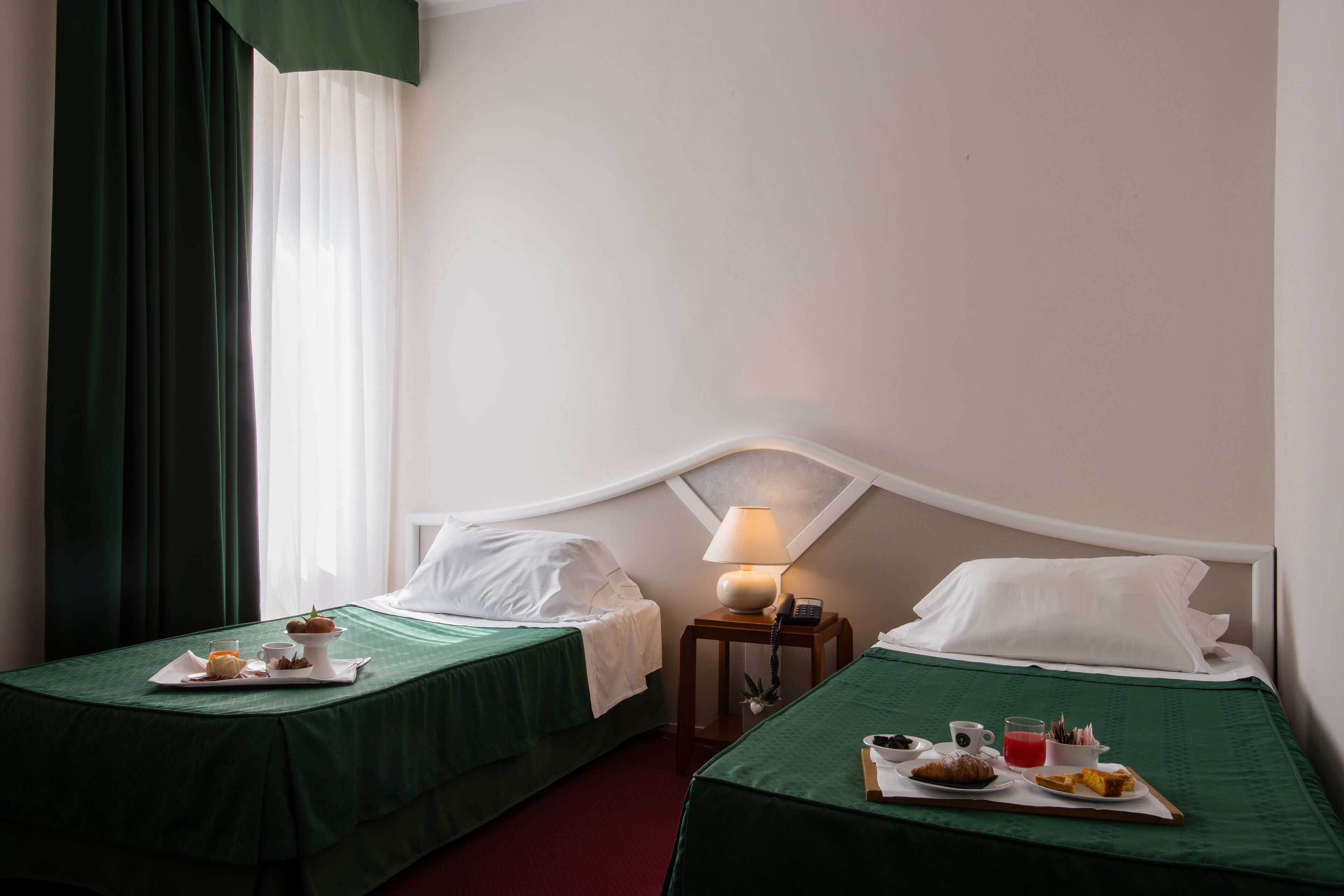 A hotel room with two beds, a nightstand with a lamp, and breakfast trays on each bed.