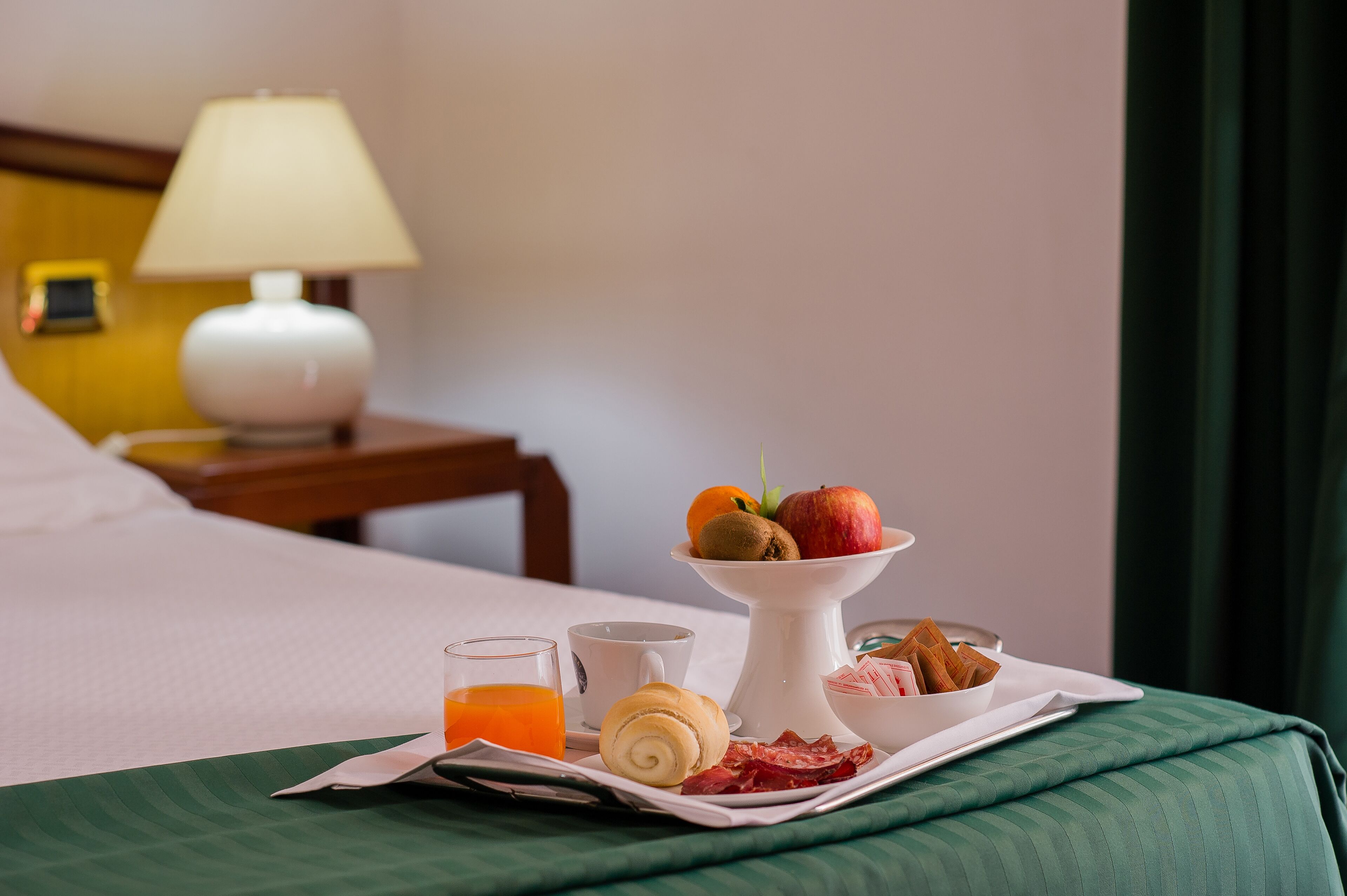 A neatly made breakfast tray with fruits, a rolled pastry, and a glass of orange juice on a bed with a green bedspread.