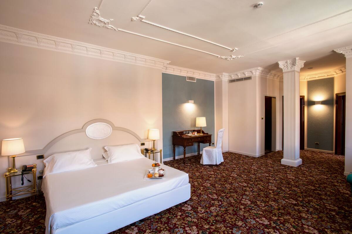 Een hotelkamer met een groot bed, twee nachtkastjes met lampen, een bureau met een stoel, en een decoratieve kolom.