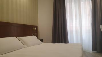 Ein Hotelzimmer mit einem großen Bett, einem Holz-Kopfteil, einem Nachttisch und einem Fenster mit transparenten Vorhängen.