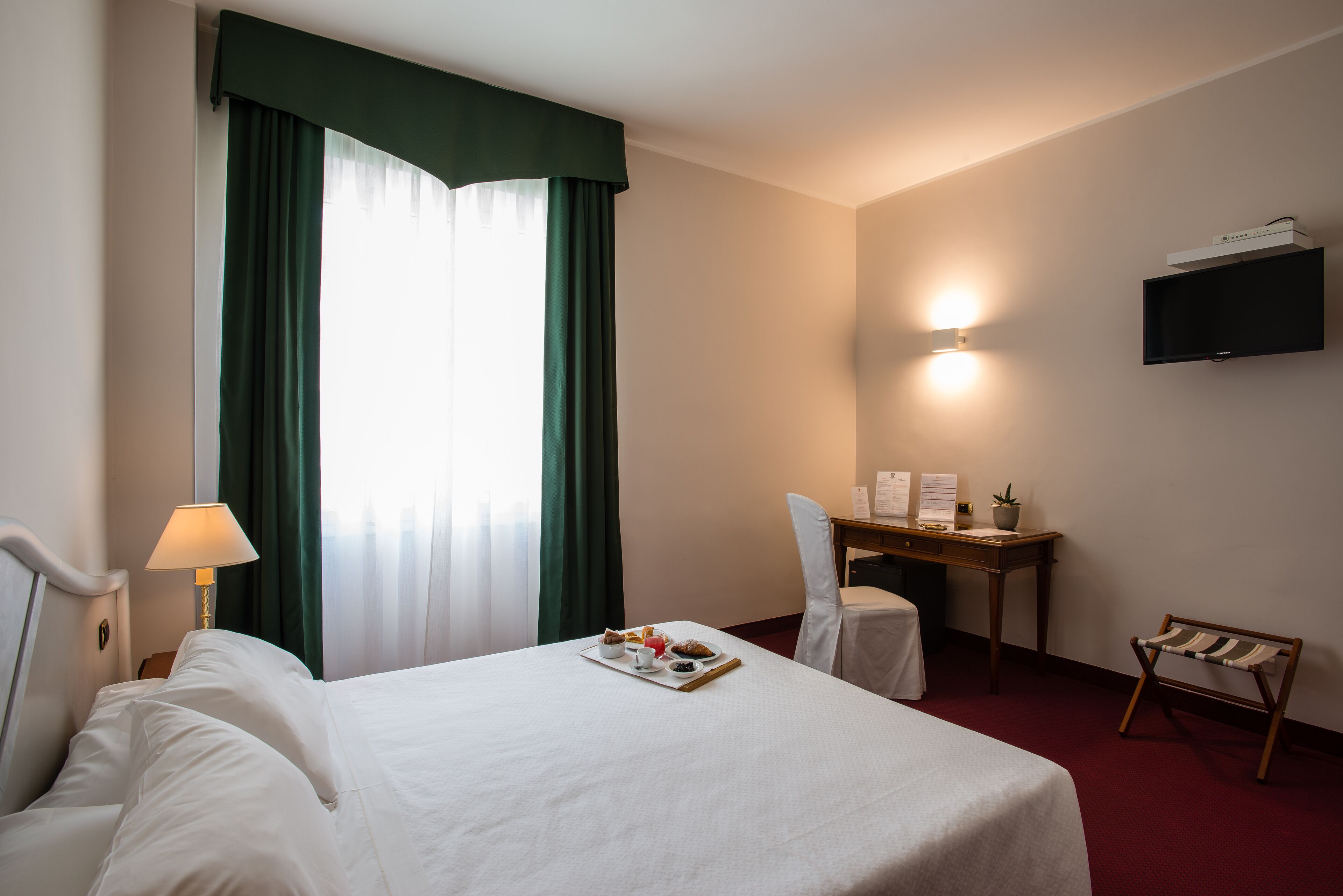 A hotel room with a bed, a desk, a chair, a TV, and a small table with a tray of breakfast items.