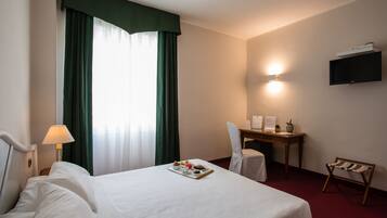 A hotel room with a bed, a desk, a chair, a TV, and a small table with a tray of breakfast items.