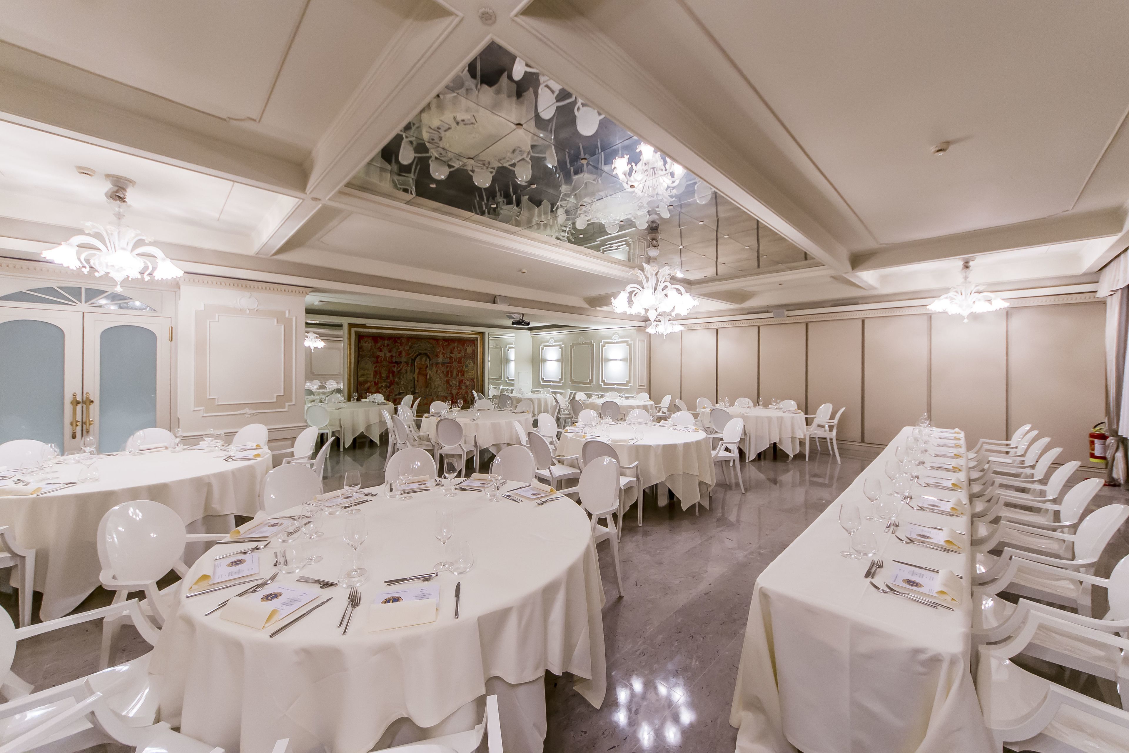 Une salle de banquet avec de nombreuses tables rondes dressées pour le repas, des chaises blanches et des lustres.