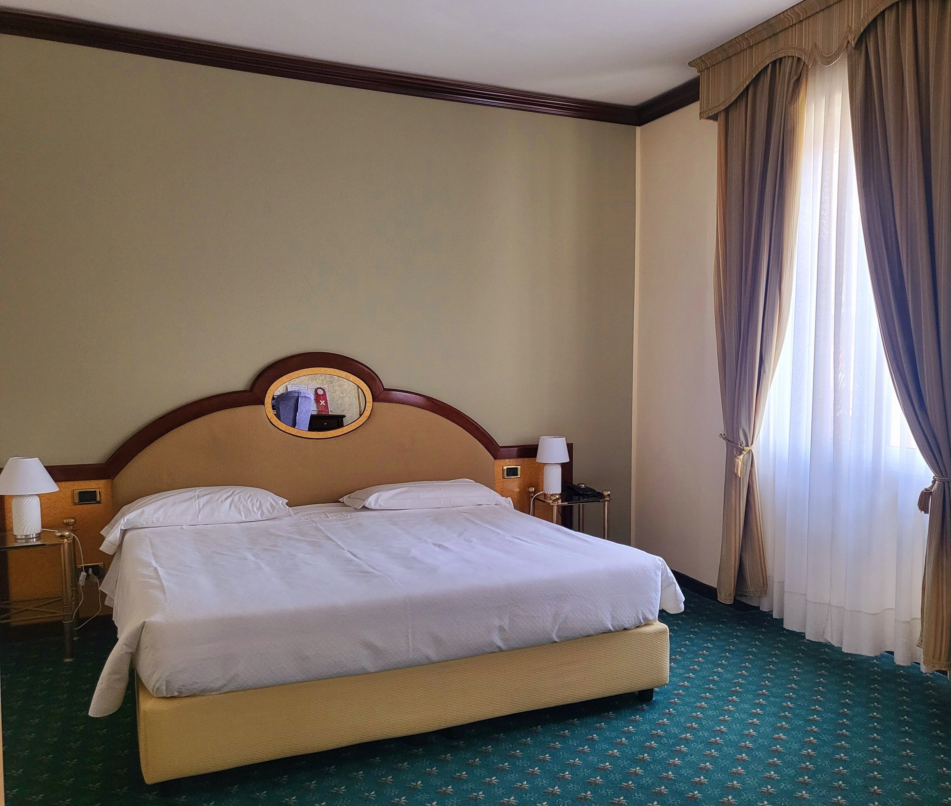 Una habitación de hotel con una cama grande, un espejo redondo sobre la cabecera, dos lámparas de noche y una ventana con cortinas.