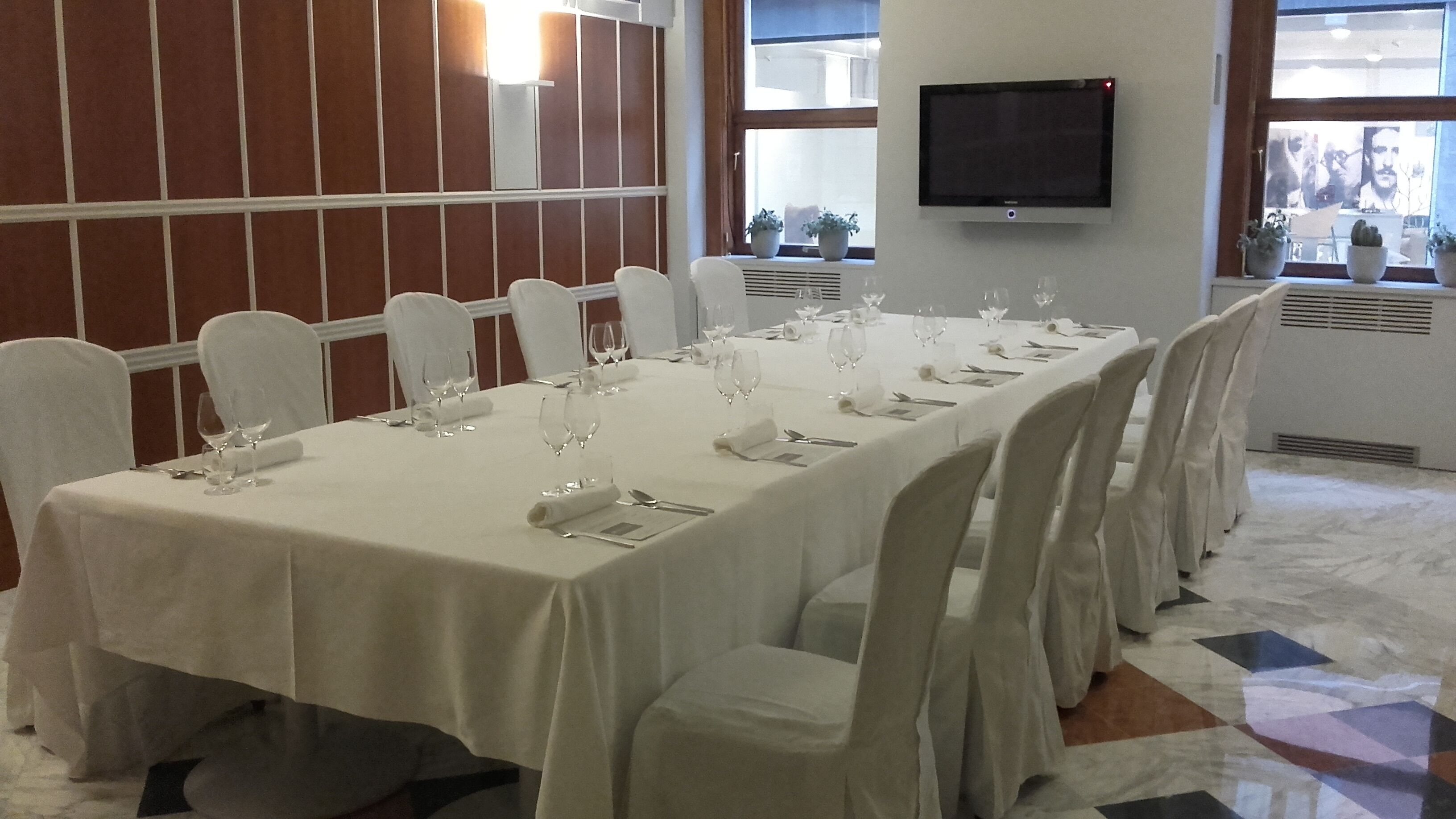 Ein Konferenzraum mit langem Tisch, gedeckten Plätzen, weißen Stühlen und einem an der Wand montierten Flachbildfernseher.
