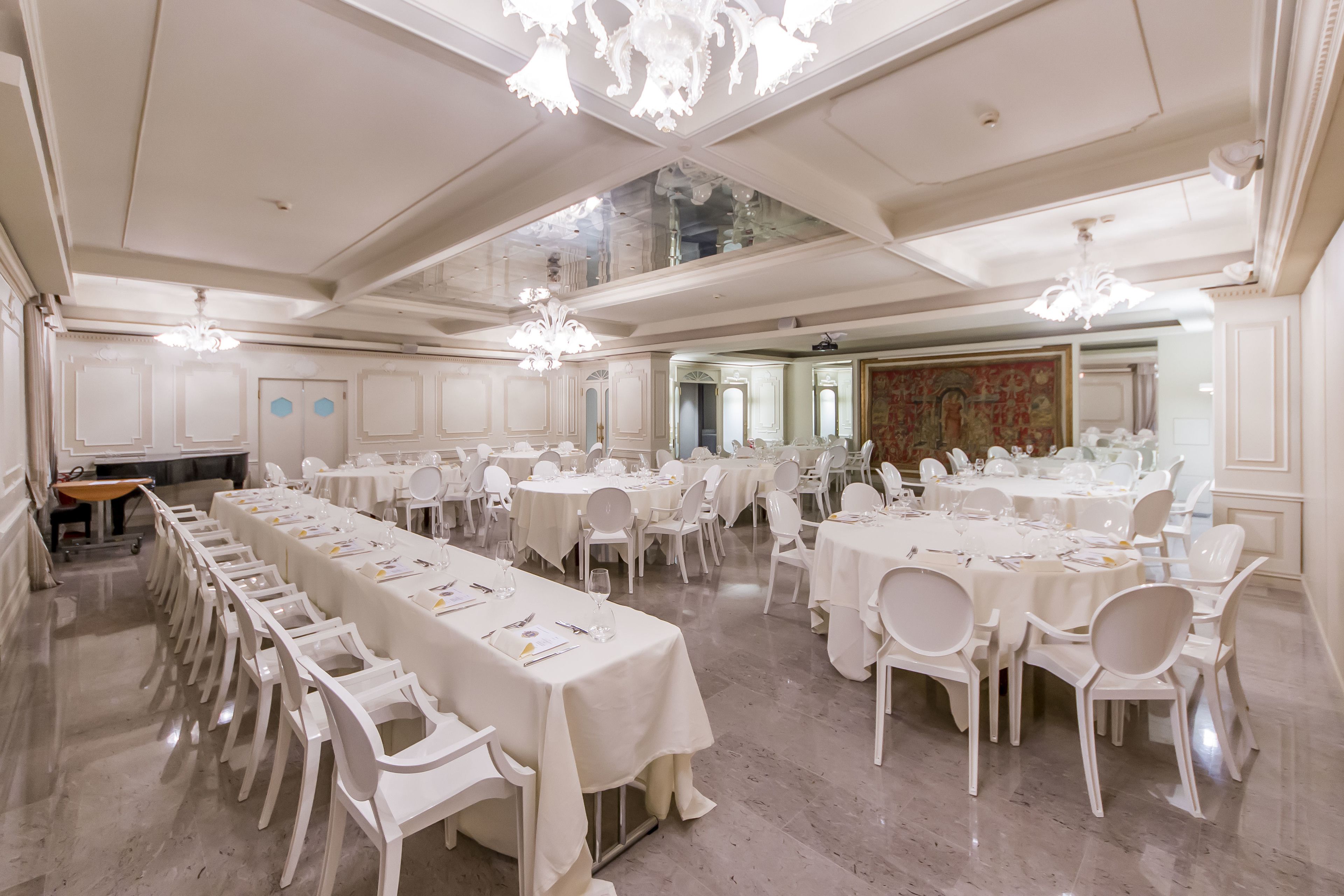 Une vaste salle de banquet dotée de longues tables et de chaises blanches, de hauts plafonds ornés de lustres somptueux et d’un sol en marbre poli.
