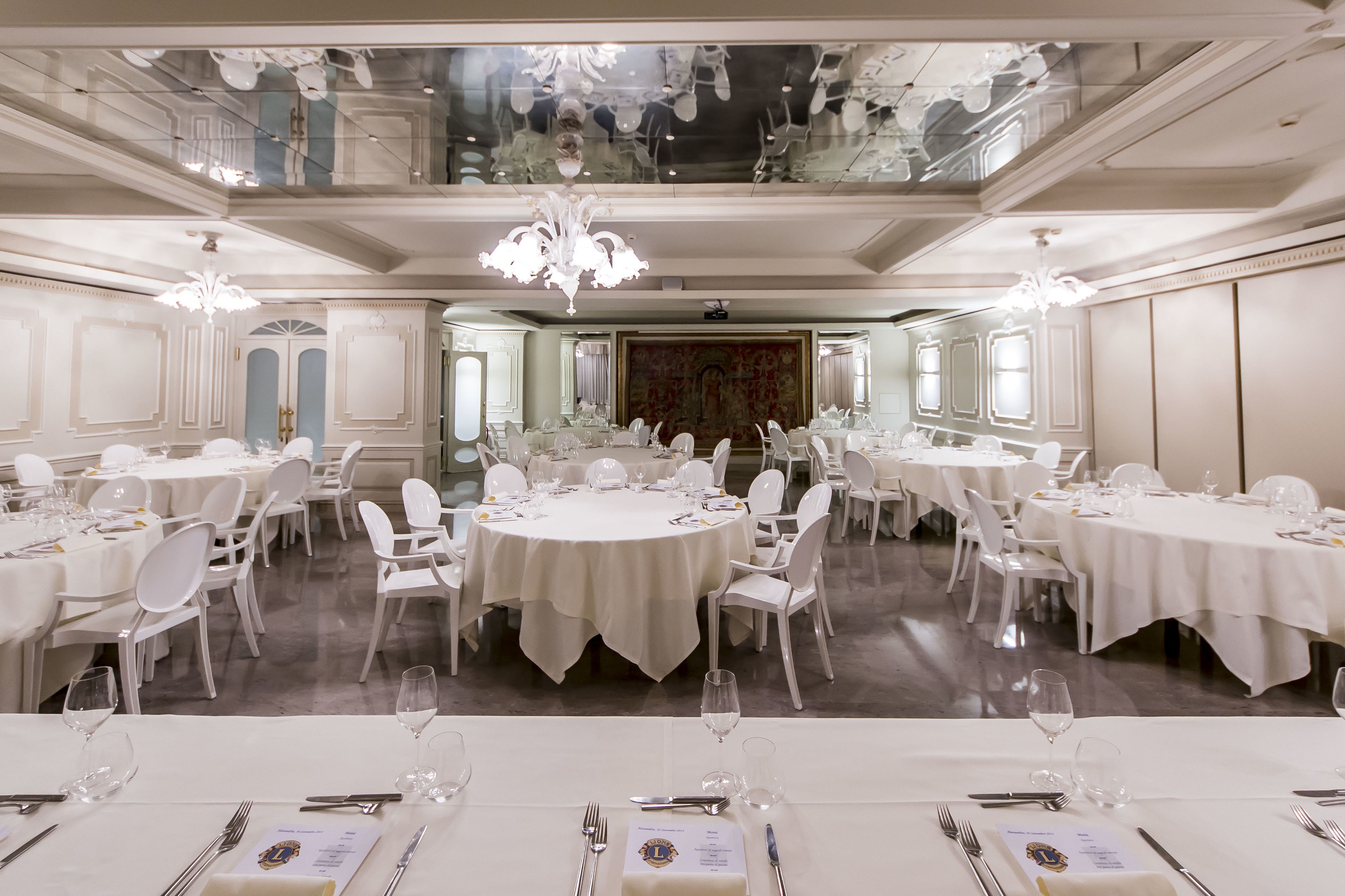 A formal dining room with multiple round tables, white chairs, and chandeliers.