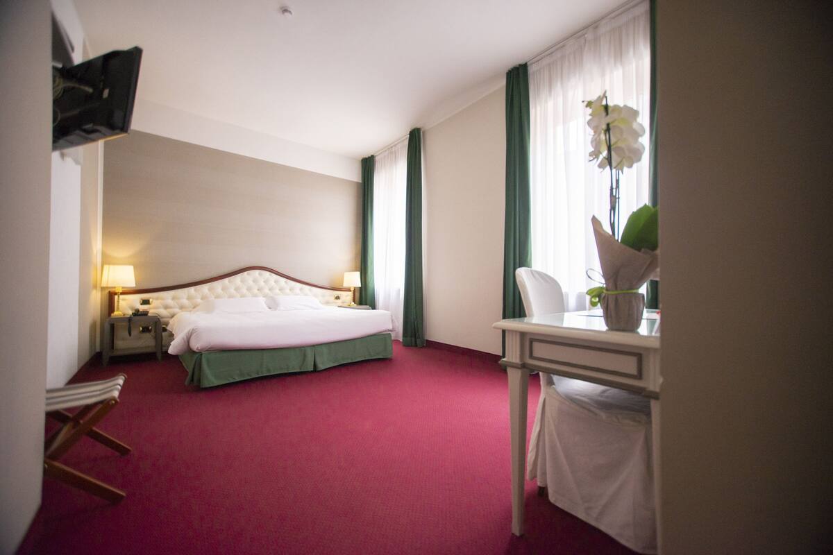 Een hotelkamer met een groot bed, een bureau met stoel, een lamp en een raam met gordijnen.