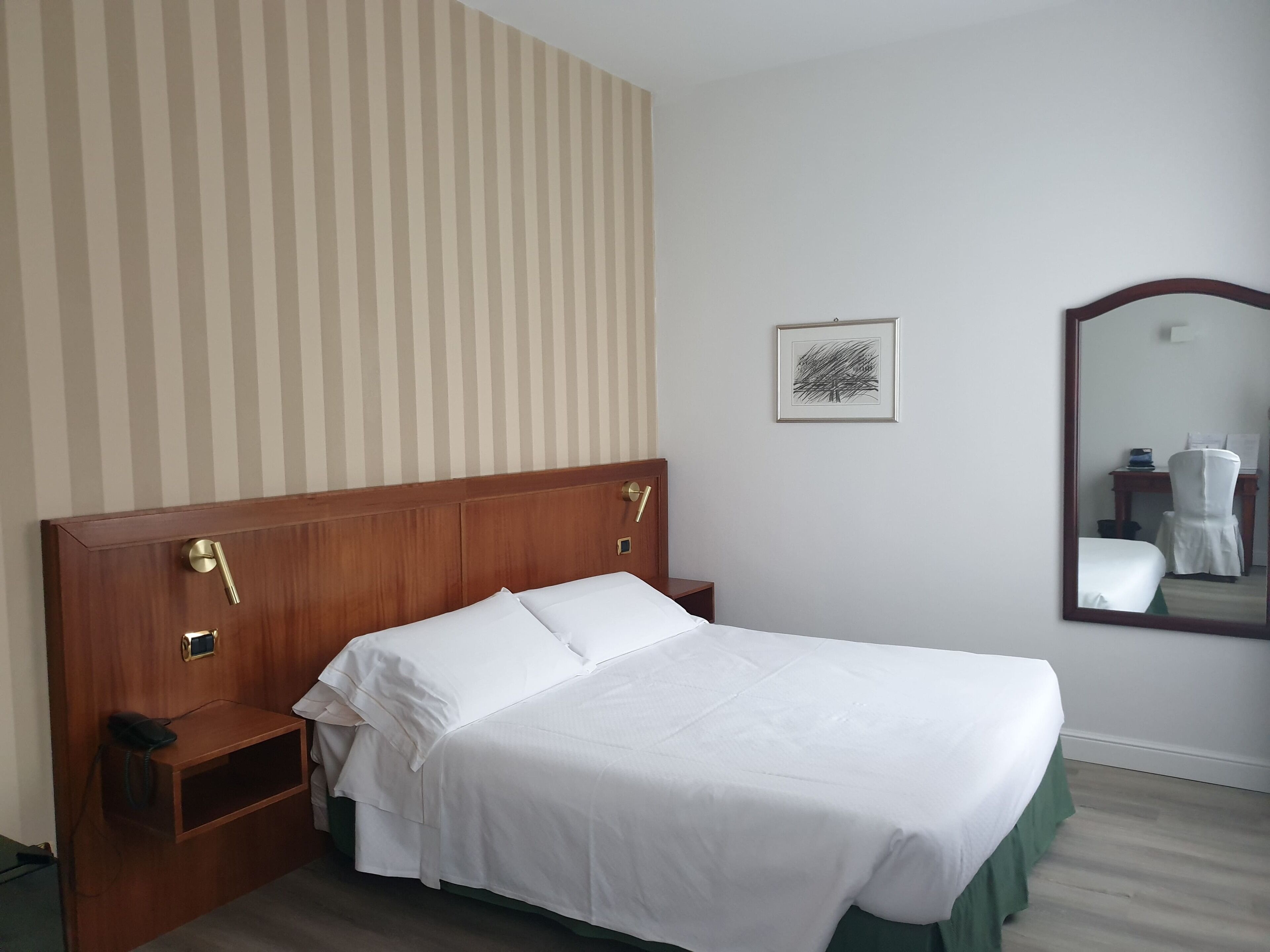 Una cama bien hecha con sábanas blancas, un cabecero de madera, una mesita de noche con teléfono y un cuadro en la pared.