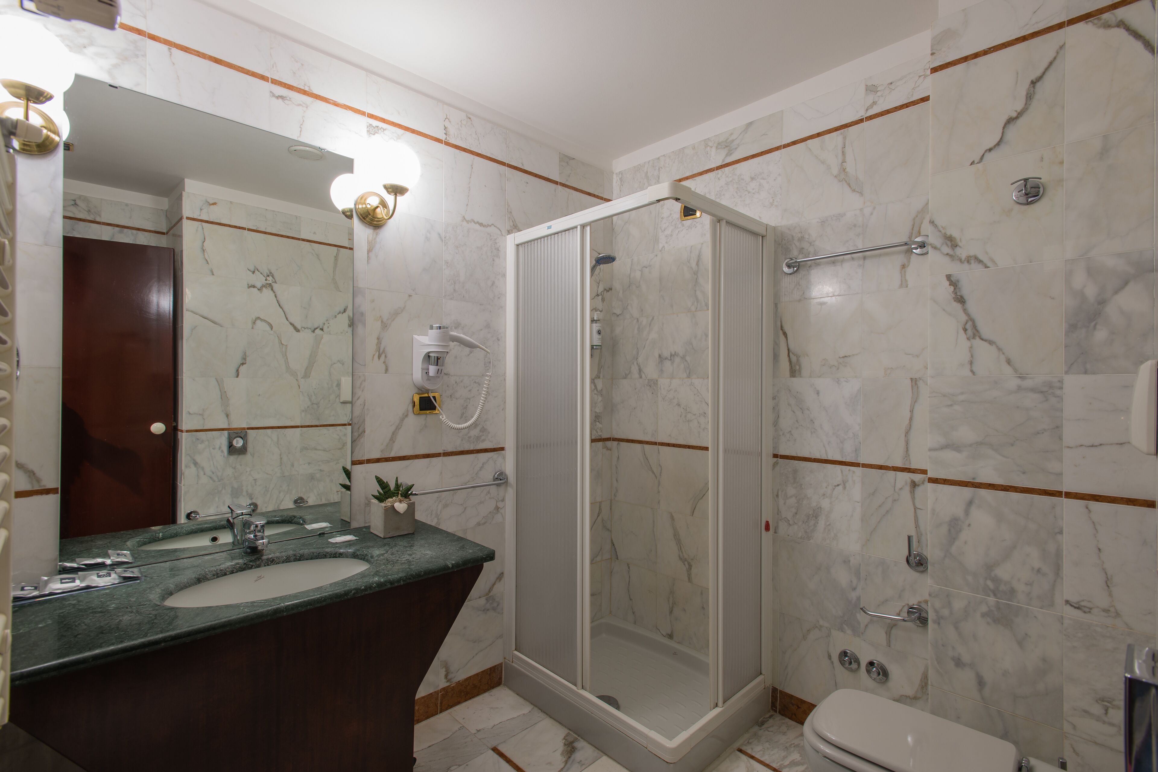 A bathroom with a marble wall, a shower, a toilet, and a sink with a mirror.
