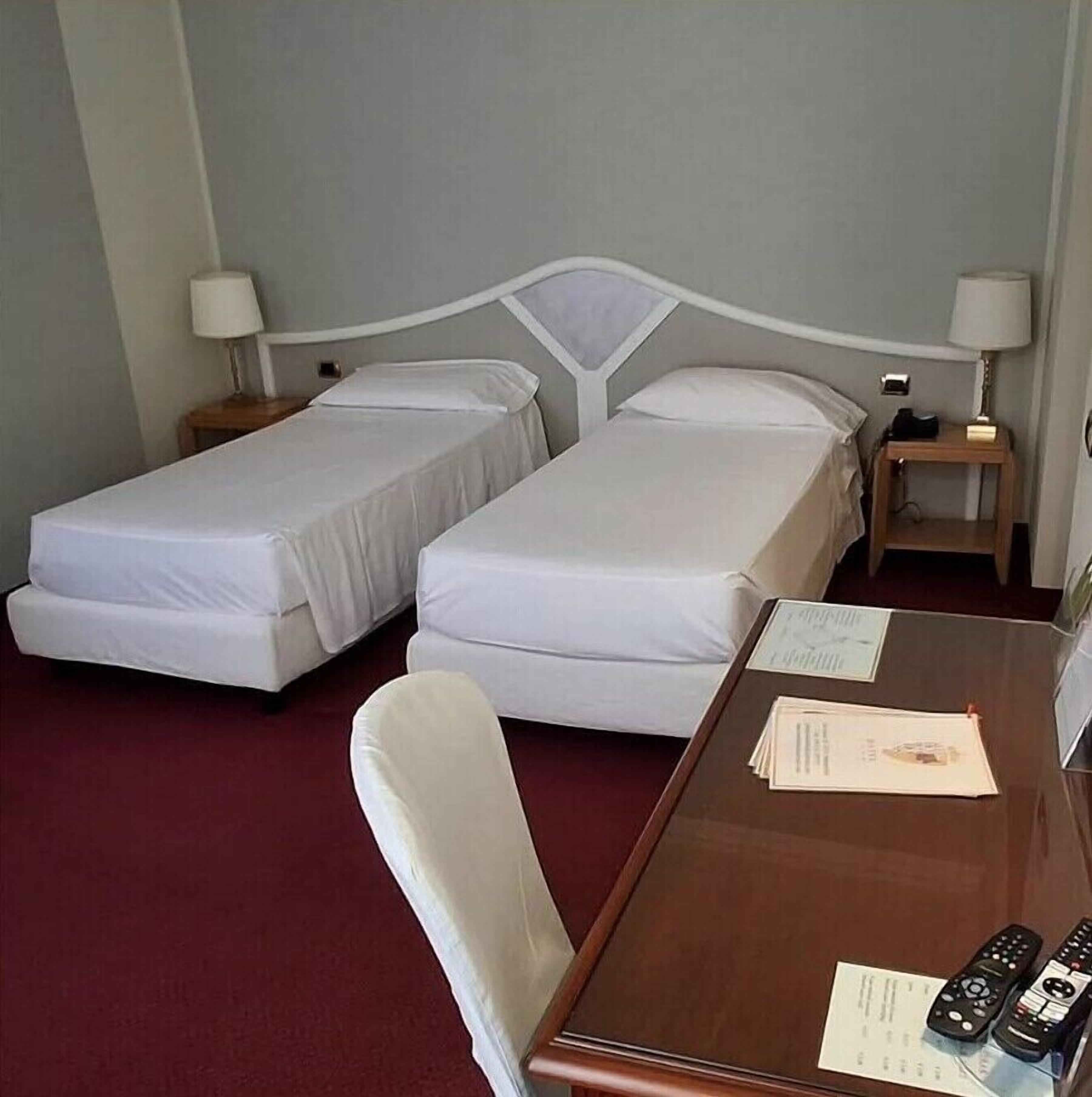 Een hotelkamer met twee eenpersoonsbedden, een houten bureau met stoel, en een nachtkastje met lamp.
