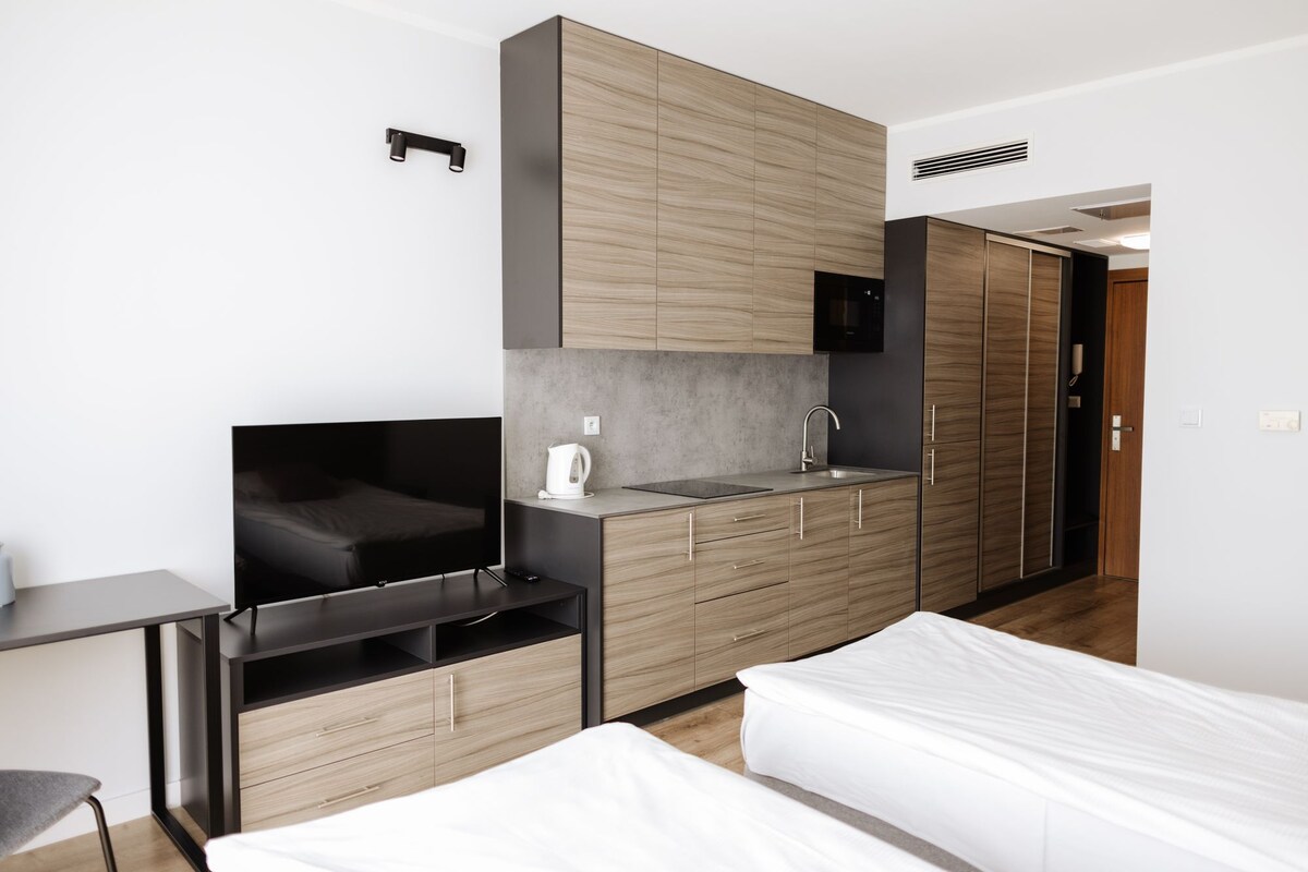 A modern hotel room with a bed, a TV, a kitchenette, and a wardrobe.