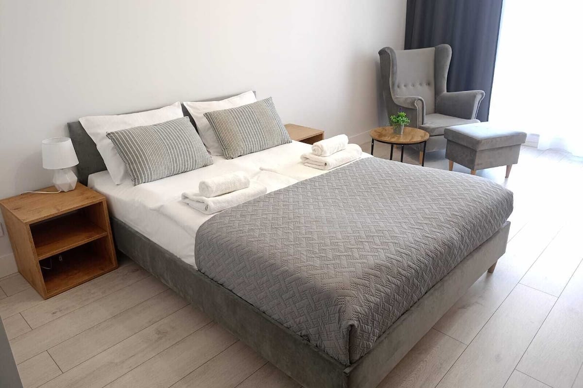 A neatly made bed with a grey quilt, two pillows, and a bedside table with a lamp.