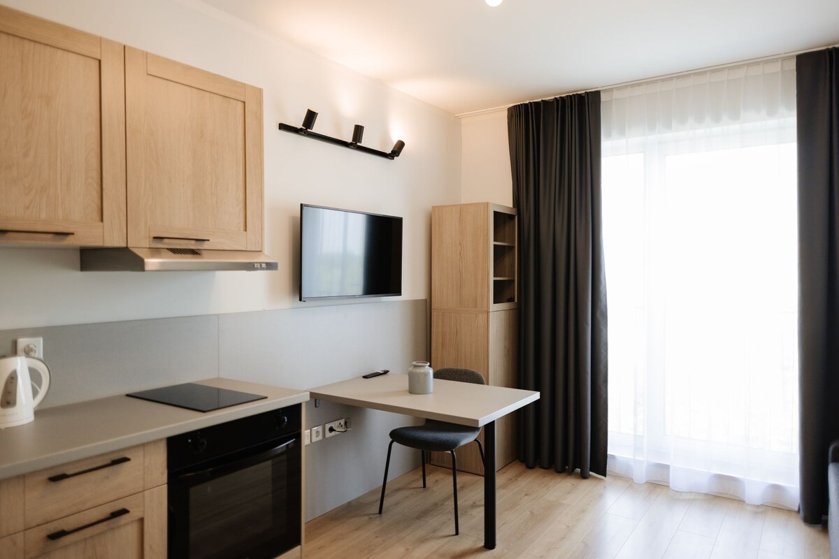 A modern kitchen with wooden cabinets, a built-in oven, a black countertop, and a dining area with a table and chair.